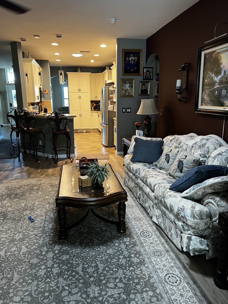 Living room with floral sofa, decorative pillows, and a wooden coffee table. A kitchen is visible in the background with white cabinets and hanging lights. Framed pictures and a large painting decorate the wall.