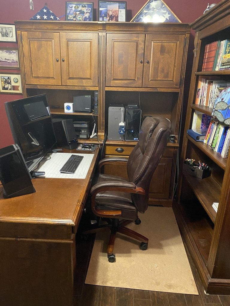 Home office with wooden desk, computer setup, leather chair, shelves with books and decorations, and framed memorabilia on the wall.