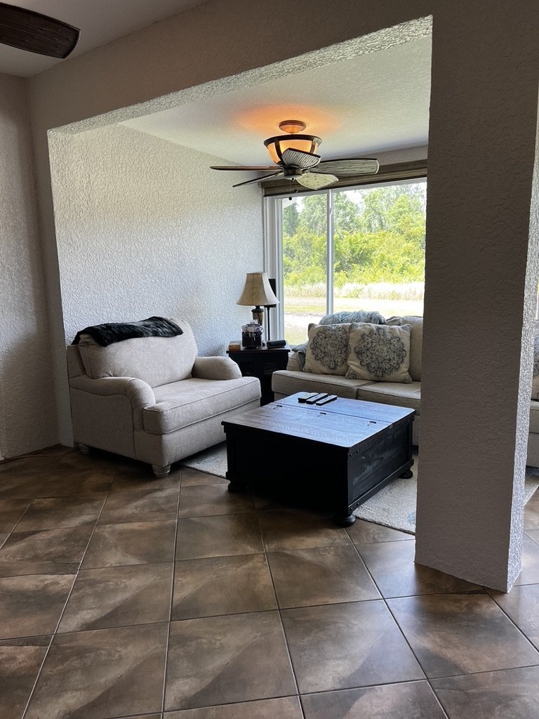 Cozy living room with armchair, sofa, coffee table, lamp, and ceiling fan, tiled floor, and large window with outdoor view.