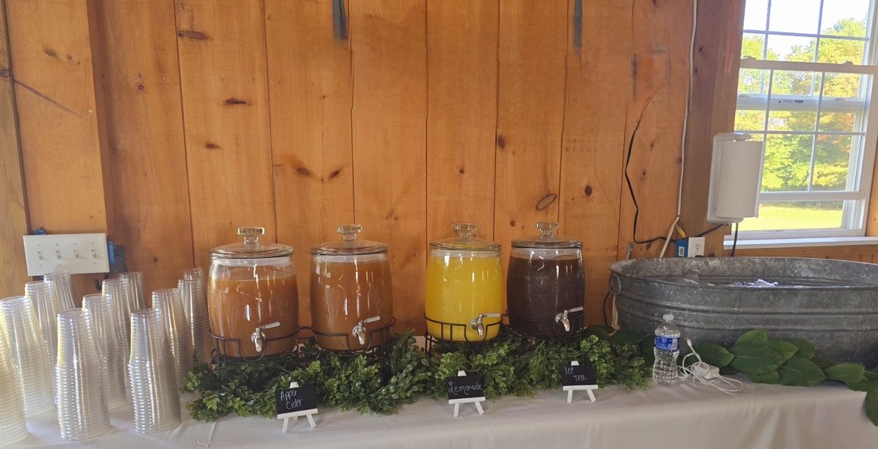 Beverage station with dispensers of apple cider, lemonade, iced tea, plastic cups, and metal tub on a table with white cloth and greenery.