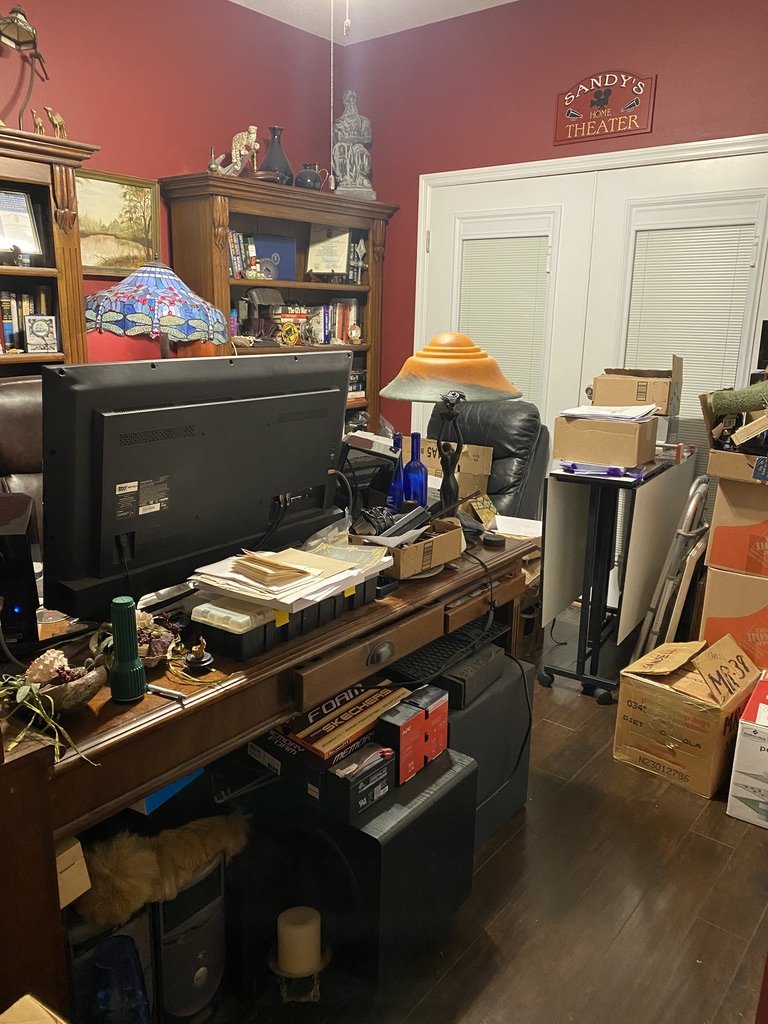 Cluttered home office with a red wall, desk with computer, lamp, and various items, bookshelves with books and decorations, boxes, and a sign labeled "Sandy's Home Theater."