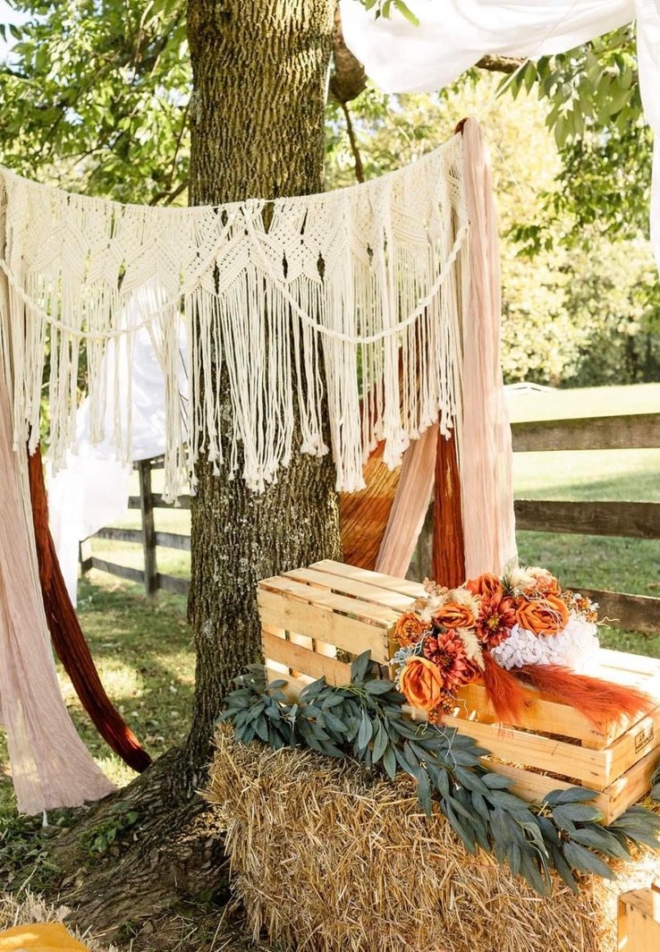 Outdoor rustic wedding decor featuring a macramé backdrop hanging from a tree, a wooden crate filled with orange and pink flowers, and greenery on a hay bale.