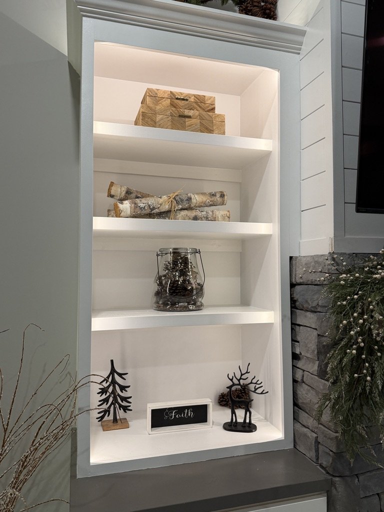 Decorative shelf with wooden boxes, birch logs, a jar with pinecones, metal tree and deer figures, a small sign reading 'Faith,' and greenery.