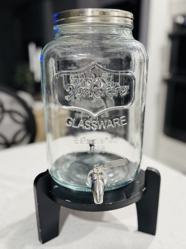 Glass beverage dispenser on a black stand with metal lid and spigot.