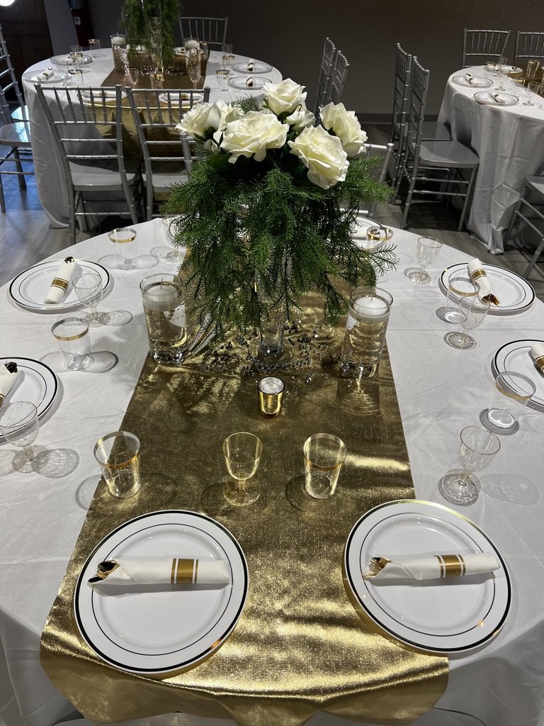 Elegant table setting with white plates and gold-trimmed glassware on a gold table runner. Centerpiece features white roses and greenery. Napkins wrapped in gold rings are placed on plates.