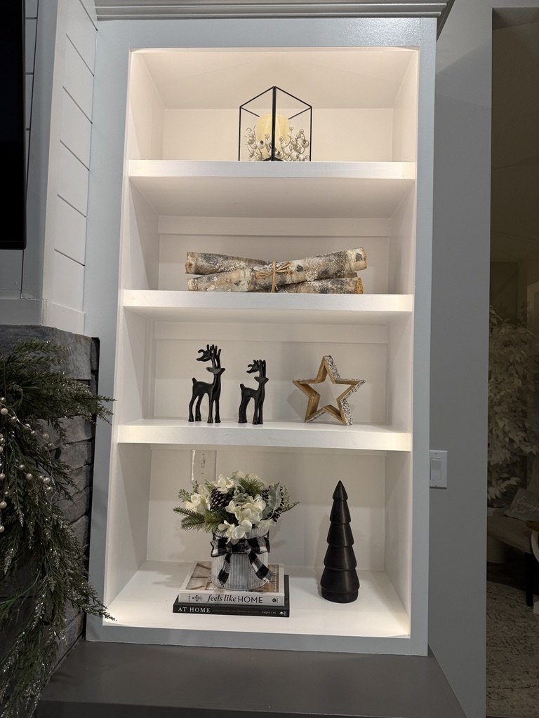 White bookshelf with holiday decor including logs, reindeer figurines, a star ornament, candle in a lantern, floral arrangement, black tree sculpture, and books.