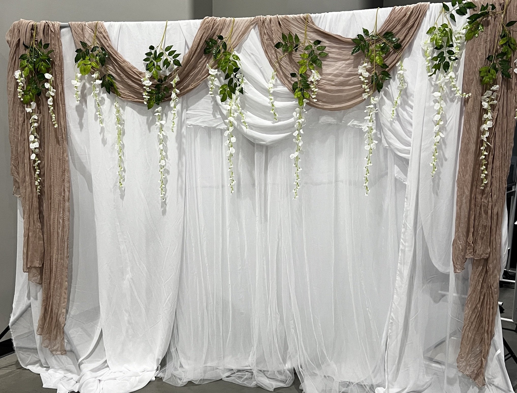 Wedding backdrop with white drapes, brown fabric, green leaves, and white hanging flowers.