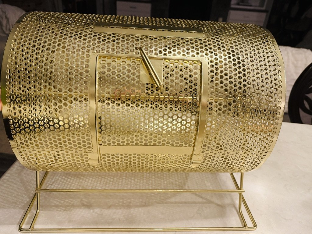 Gold-colored metal raffle drum with perforated design on a white surface.