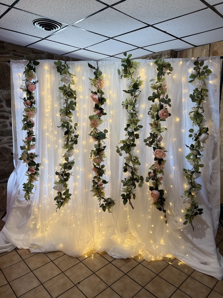 Floral and light backdrop with white drapes, green leaves, and pink flowers, set in an interior space with a tiled floor and ceiling.