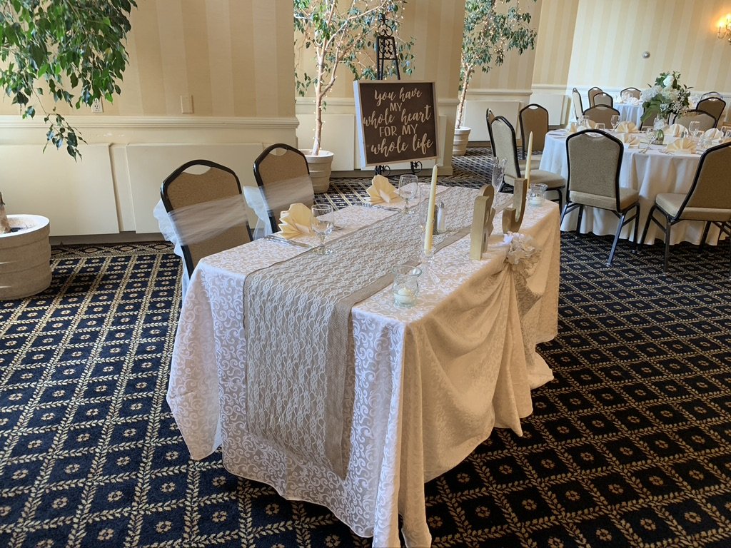 Wedding reception table setting with elegant white tablecloths, beige chairs, decorative drapes, and a sign reading "You have my whole heart for my whole life."