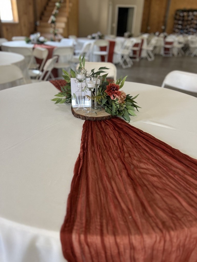 Table setup in an event venue with a white tablecloth and rust-colored table runner, adorned with candles, flowers, and greenery centerpiece on a wooden slab, surrounded by white chairs.