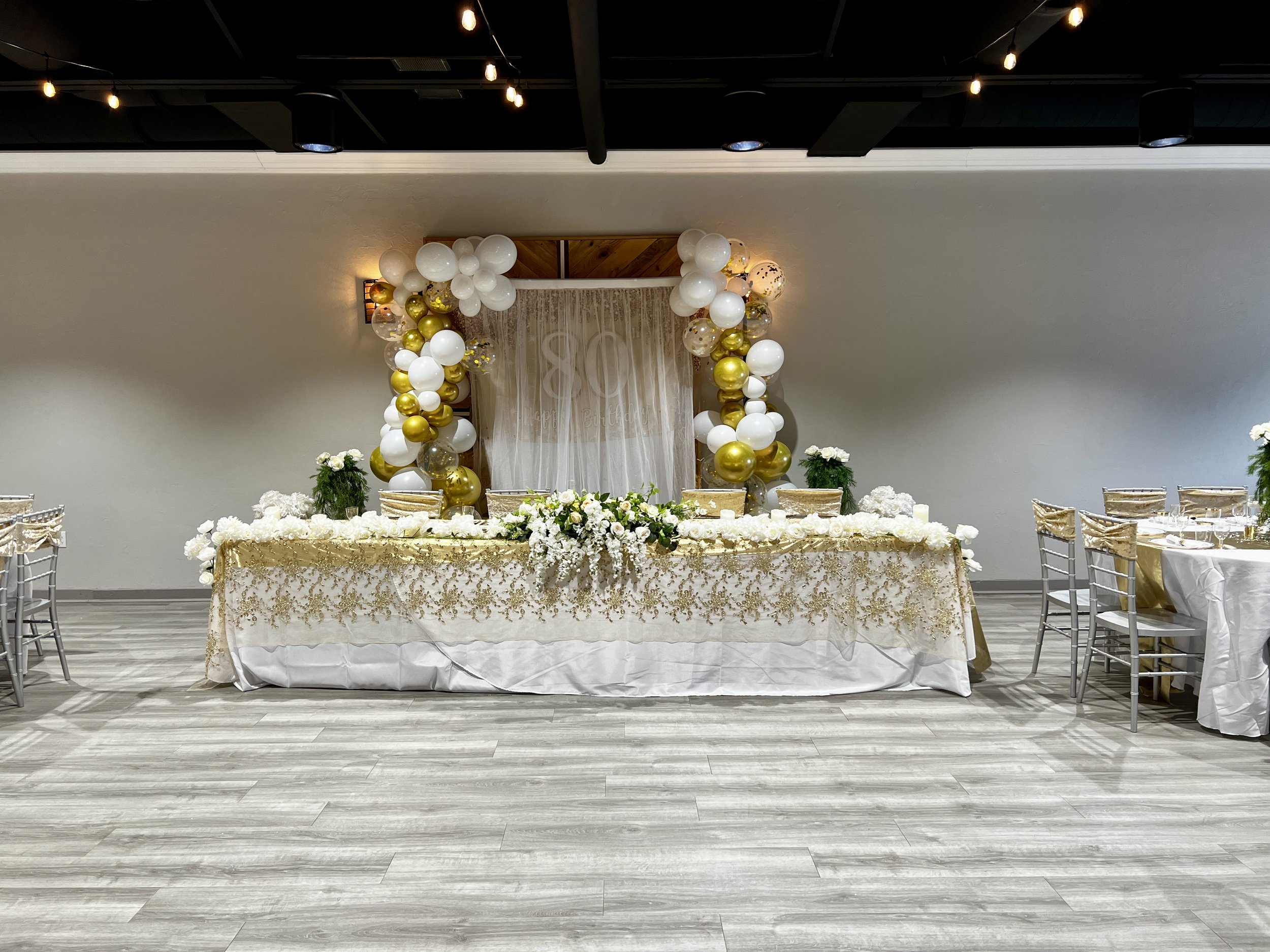 Elegant banquet table with white and gold decorations, including balloons and floral arrangements. The setting includes chairs draped with gold sashes, and a backdrop featuring a large "80" in the center.