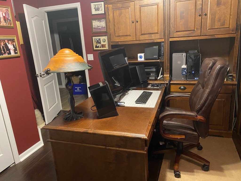 Home office setup with wooden desk, brown leather chair, computer monitors, desk lamp, and wall cabinets in a room with red walls.