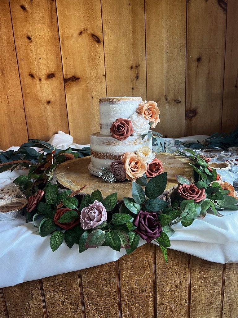 Two-tier naked cake decorated with peach and white roses, surrounded by greenery on a wooden board, set on a white cloth.