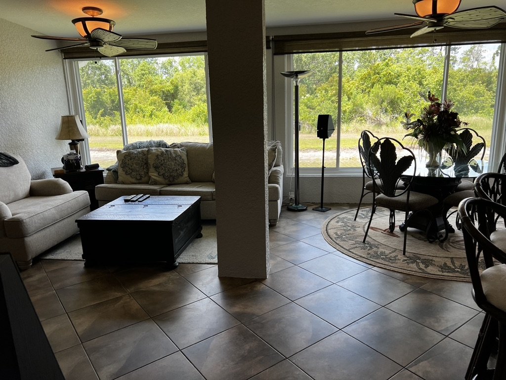 Living room with large windows, beige sofas, dark wooden coffee table, ceiling fans, and a dining area with glass table and floral centerpiece.