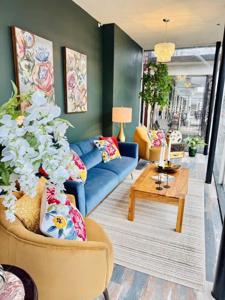 Modern living room with a blue sofa, yellow chairs, floral cushions, wooden coffee table, two floral paintings, and a tall green plant. The room features a dark green accent wall, decorative light fixtures, and a large window with a view outside.