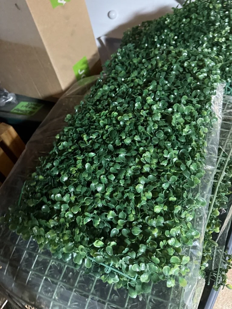 Artificial greenery resembling boxwood hedge panels, stacked on a surface, with partially visible cardboard boxes nearby.