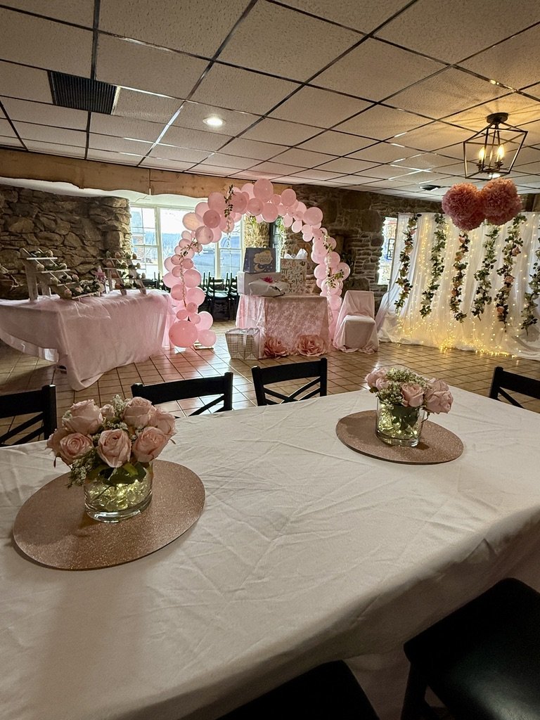 Indoor event venue decorated with a pink and white color scheme, featuring a pink balloon arch, flower arrangements, and a dessert table with cupcakes. The space includes hanging floral decorations and draped fabric with lights in the background.