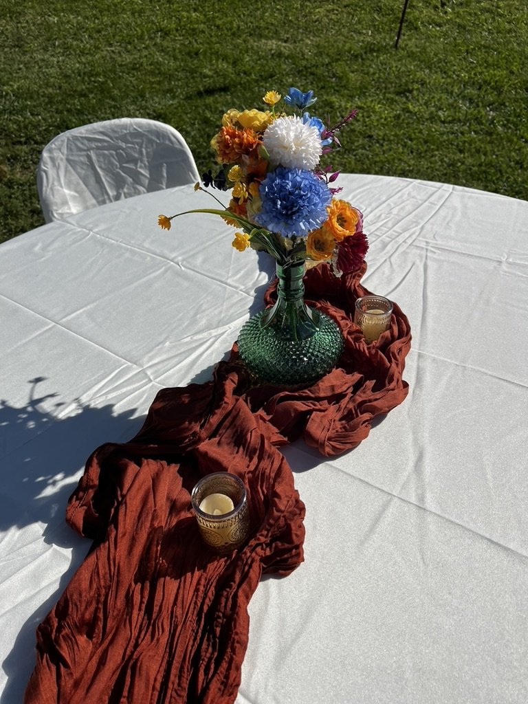 Outdoor table with white tablecloth, centerpiece of colorful flowers in a green vase, brown decorative cloth, and two small candles.