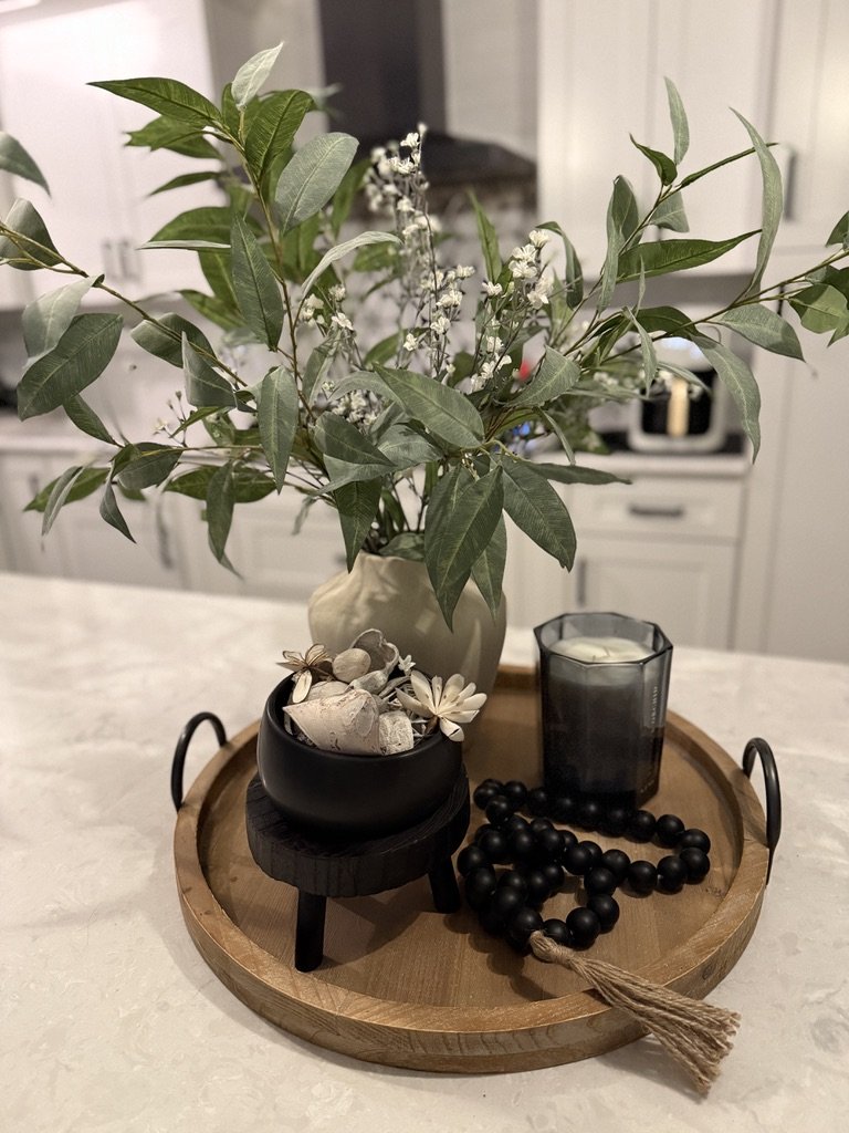 Decorative wooden tray with a vase of green leaves and white flowers, a black candle, a bowl with decorative items and flowers, and black wooden beads with a tassel on a white kitchen countertop.