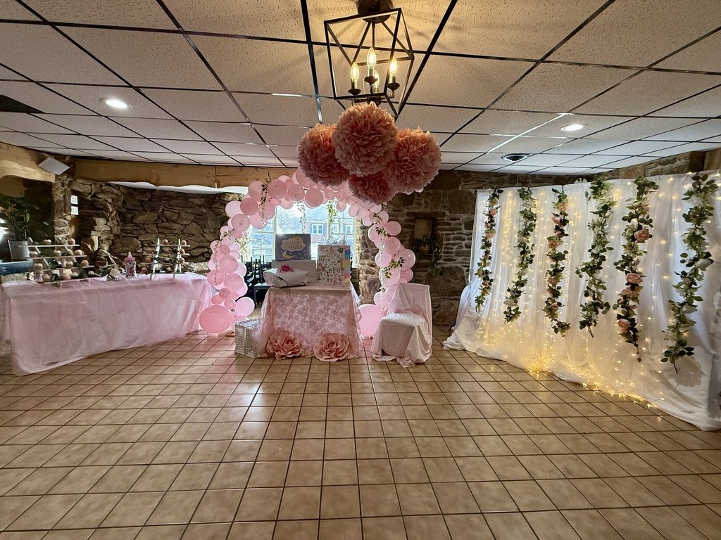 Decorated event space with pink balloon arch, floral wall, table with gift boxes, and pink tablecloths.