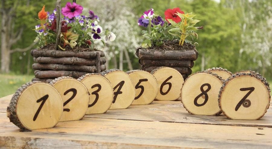 Round wooden discs numbered 1 to 8 on a table with rustic planters filled with colorful flowers in the background.