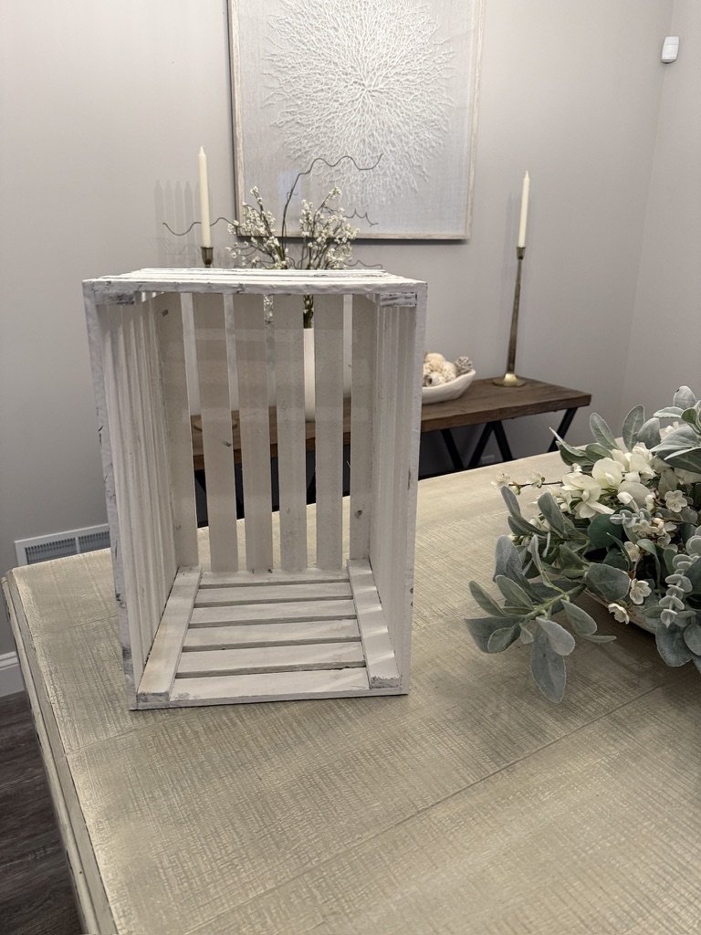 Decorative white wooden crate on a table with flowers, with a painting and candlesticks in the background.