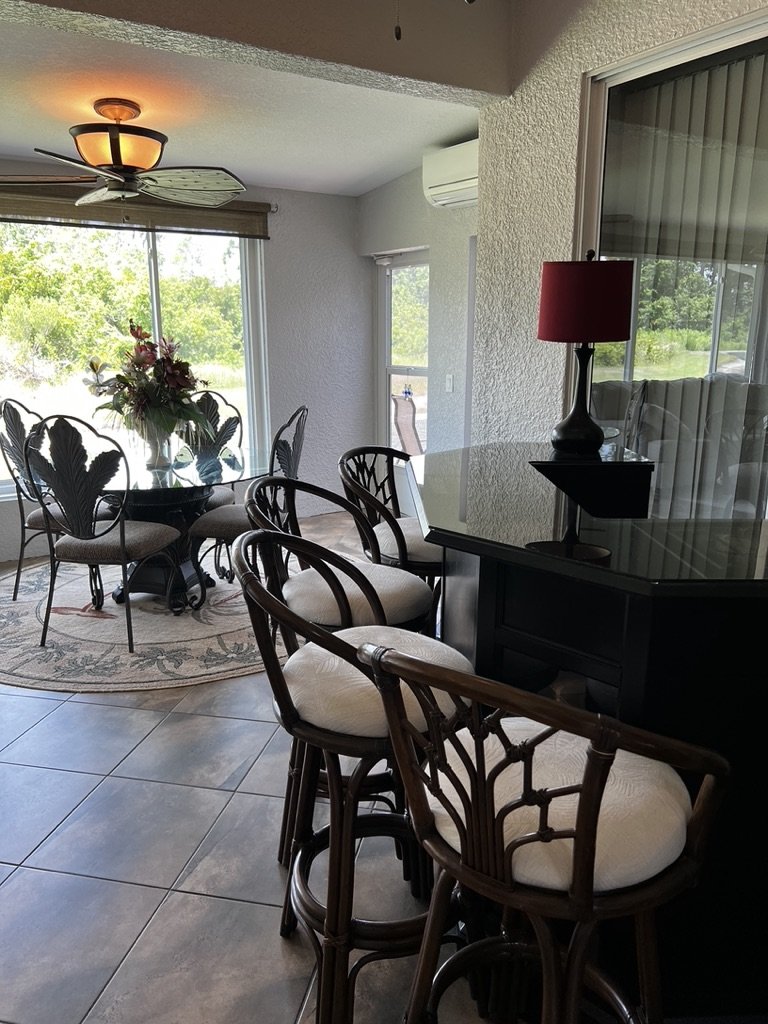 Dining area with bar stools, a round glass table, chairs, a ceiling fan, and a window view of greenery.