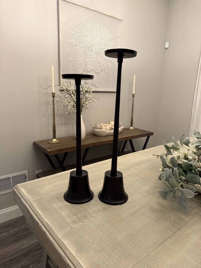 Decorative dining room setup with two black candlestick holders on a light-colored table, floral arrangement, wall art, and a console table with candles in the background.