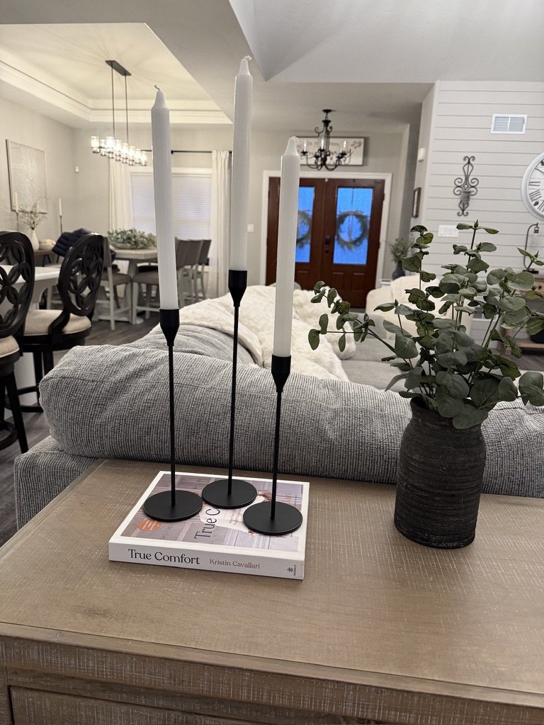 A living room scene featuring a wooden table with a book titled "True Comfort," three tall black candle holders with white candles, and a vase with green eucalyptus branches. The background shows a cozy interior with chairs, a dining table, and a cha