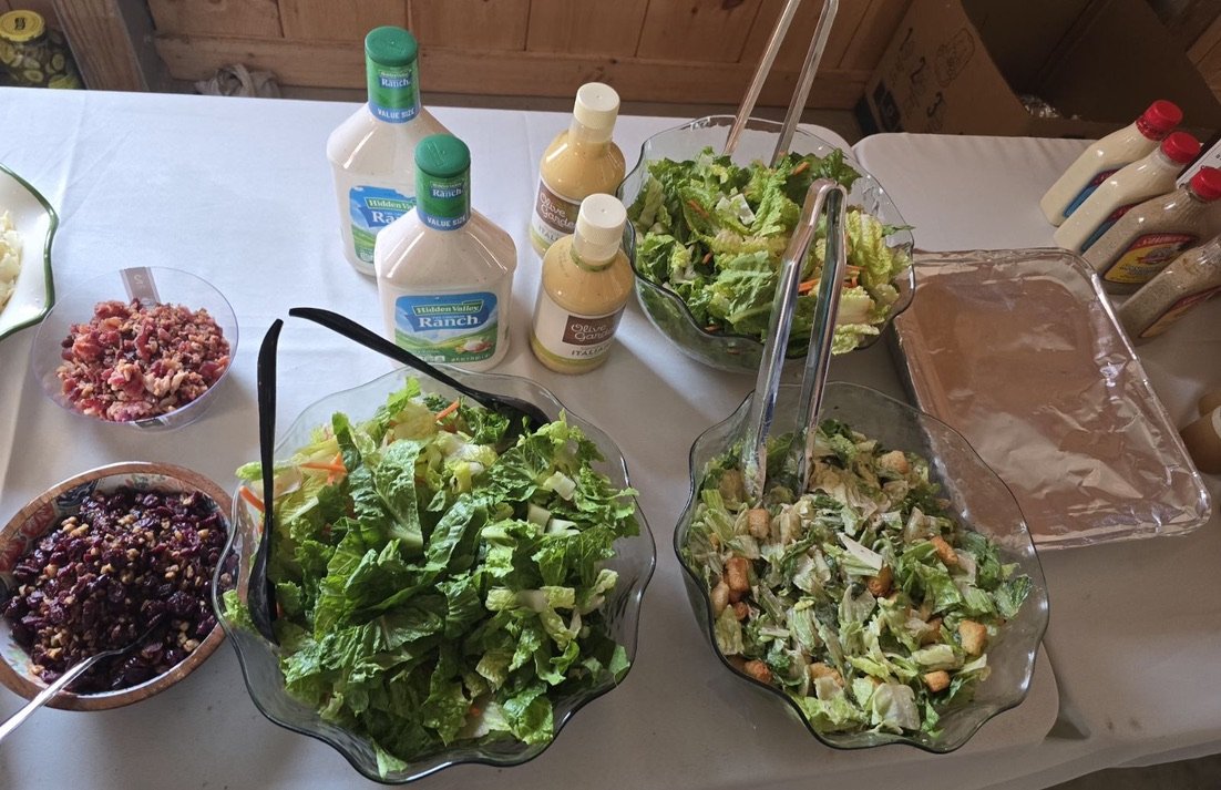 Salad bar setup with bowls of mixed greens, toppings, and bottles of ranch and caesar dressings on a table.
