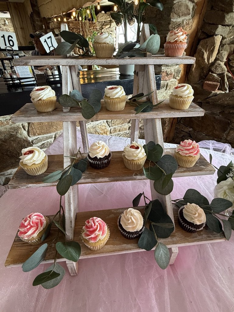 Cupcakes on a wooden tiered stand, decorated with greenery, placed on a pink tablecloth. The cupcakes have various icing designs, and a rustic stone wall is in the background. Tables numbered 16 and 17 are visible.