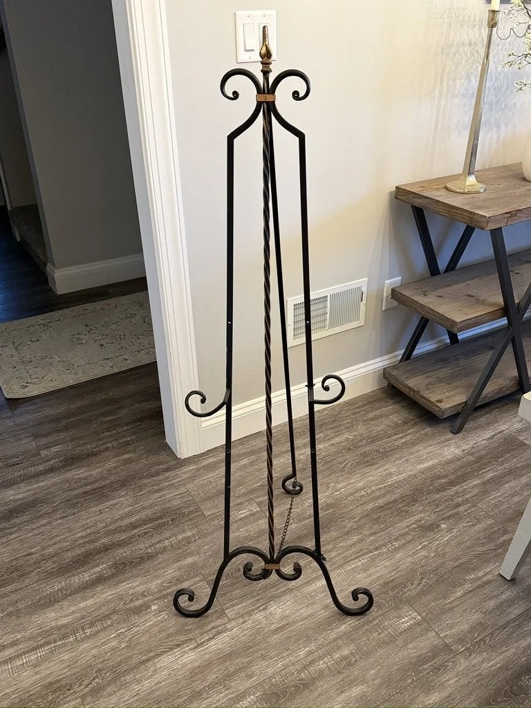 Iron easel stand for artwork or signage, located in a room with wooden floors and adjacent wooden furniture.
