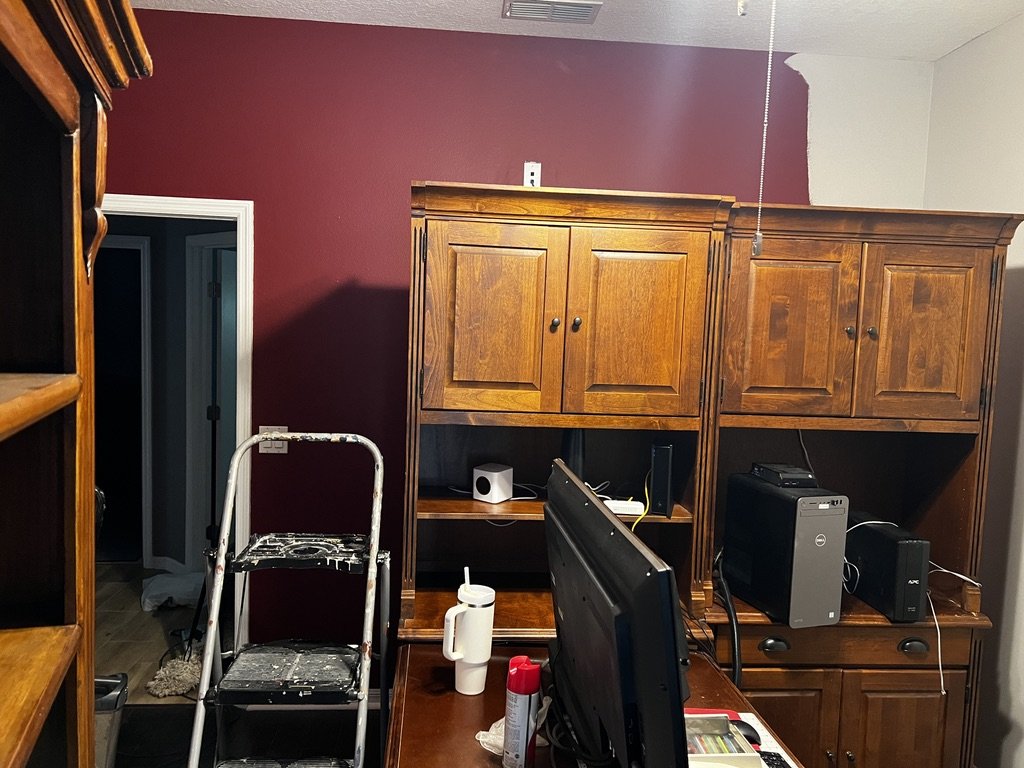 Office room with wooden furniture, including a desk and cabinets, and an unfinished maroon and white painted wall. A ladder covered in paint is near the doorway, and computer equipment is visible on the desk.