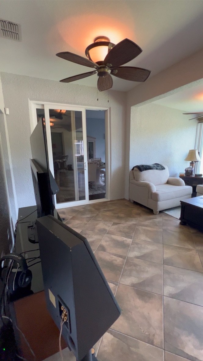 Interior room with ceiling fan, sliding glass doors, tile flooring, television on a stand, beige armchair, side table with lamp, and a cabinet.