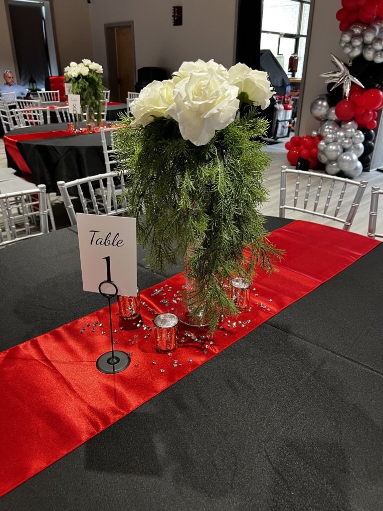Table setting for an event with a centerpiece of white roses and greenery. Red and black tablecloths with a red runner. Numbered table card "Table 1" and small decorative candles.