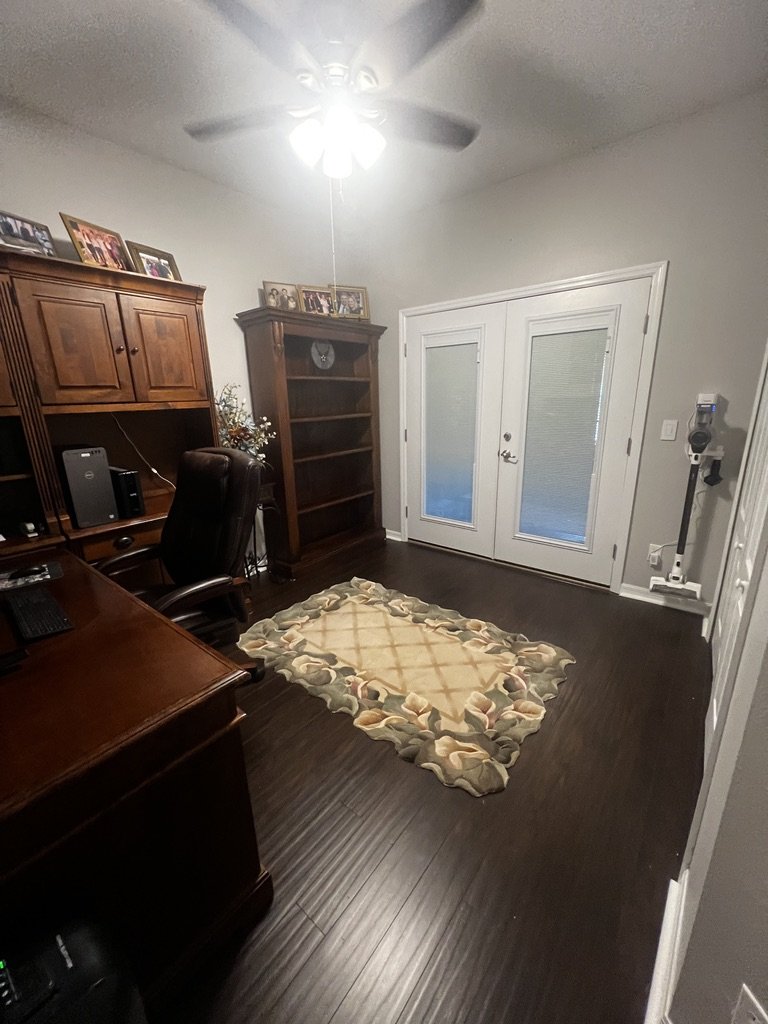 Home office with a wooden desk, office chair, and computer setup. A rug is on the dark hardwood floor. Shelves with decorative items and photos are present. There are double glass doors and a ceiling fan.