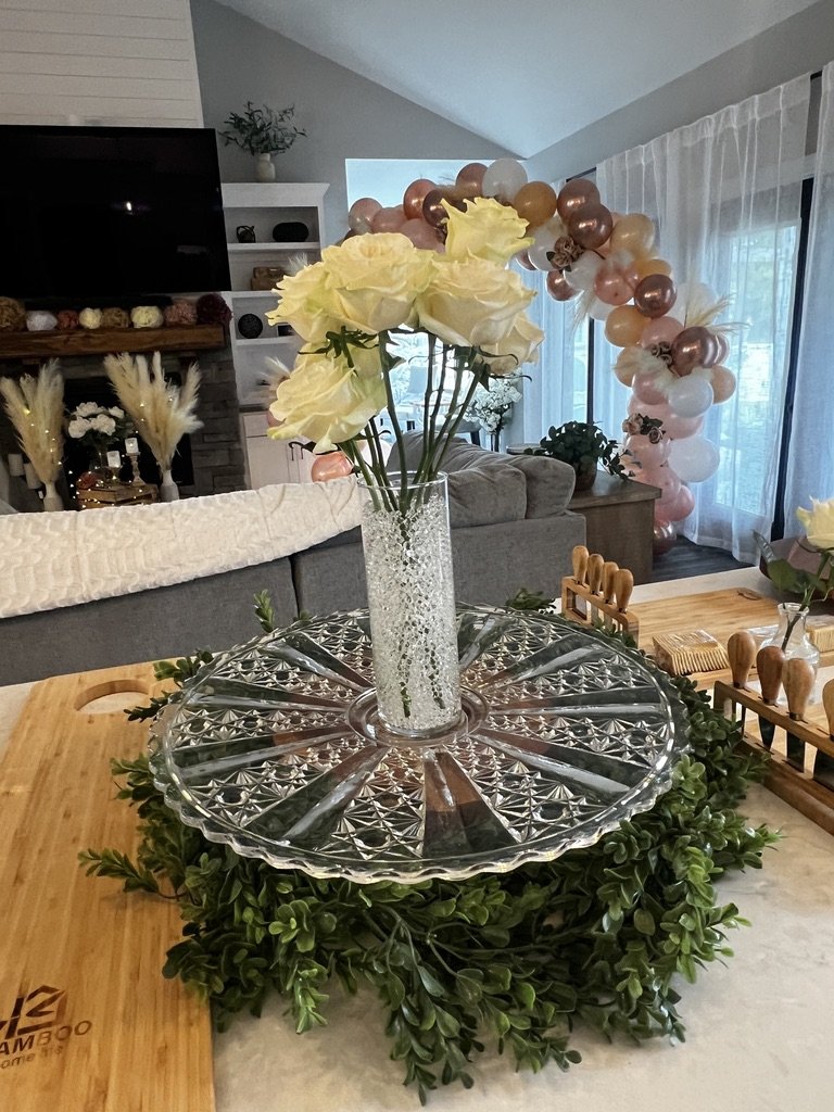 Interior room with a vase of white roses on a glass platter surrounded by green foliage, bamboo cutting board, and decorative balloon arrangement in the background.