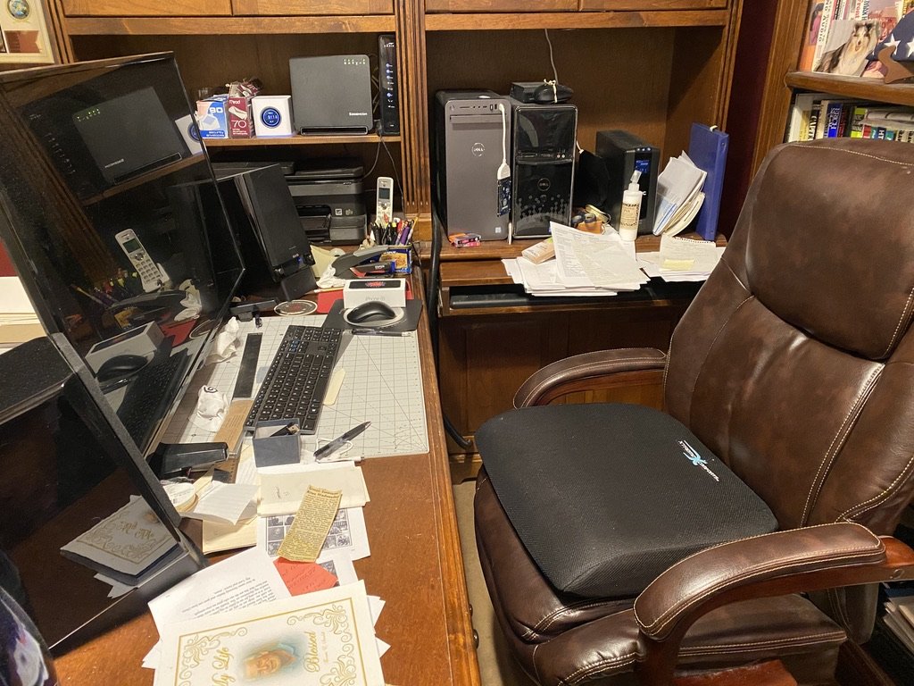 Home office setup with a cluttered wooden desk, computer monitors, keyboard, office supplies, paperwork, and a brown leather office chair. Shelves with books are in the background.