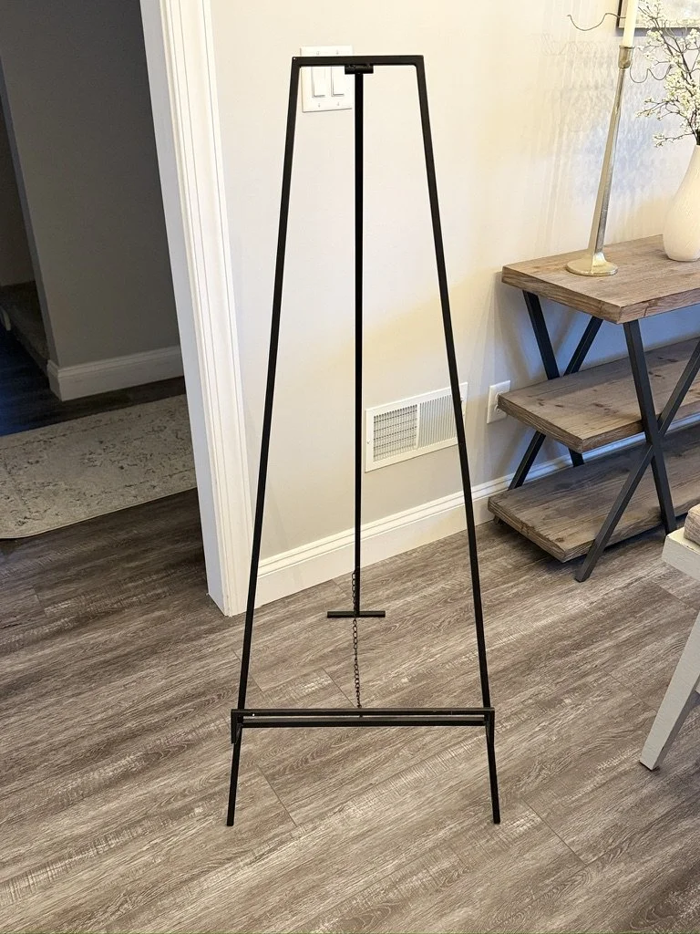 Black metal easel in a room with wooden floors and light-colored walls.