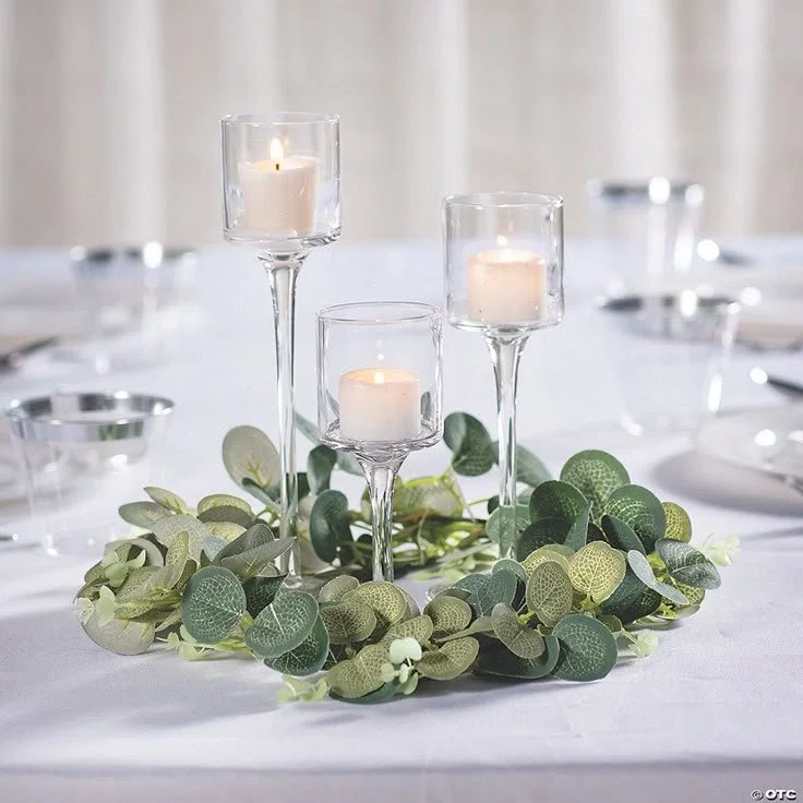 Elegant table centerpiece with three glass candle holders, lit candles, and a green leaf garland on a white tablecloth.