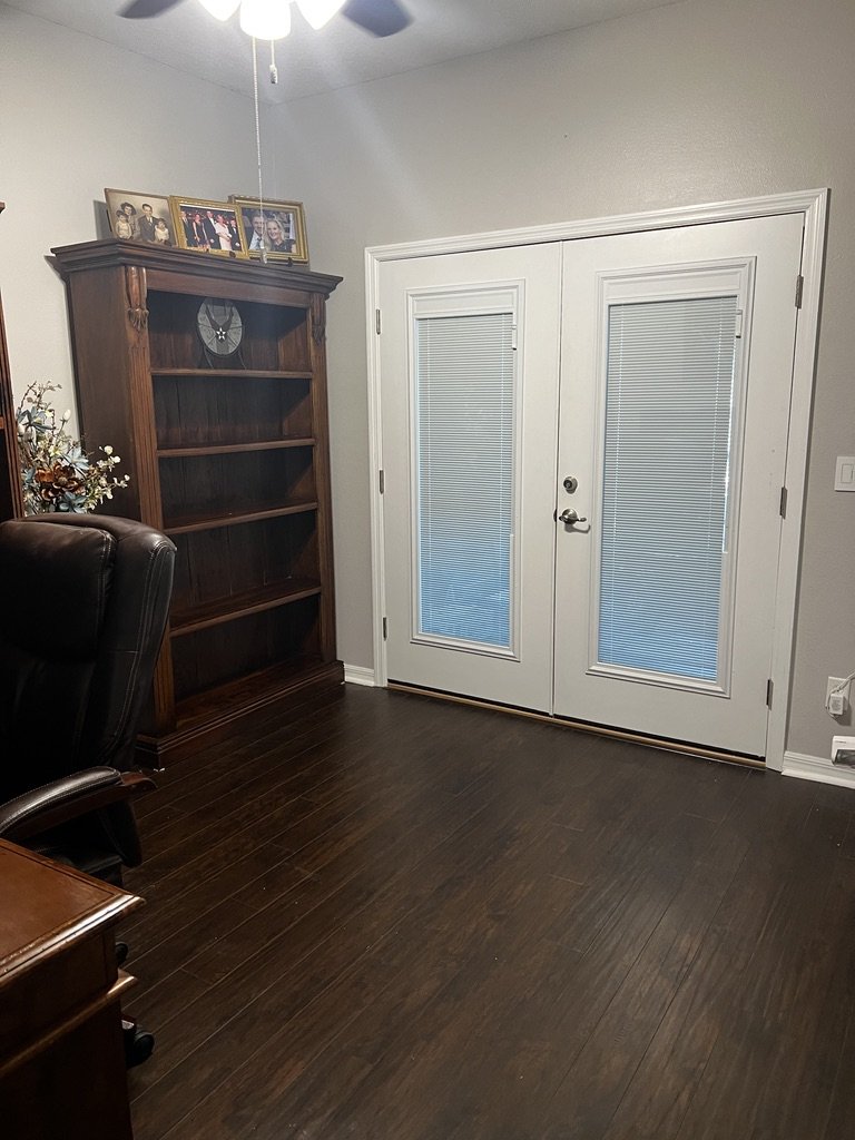 Room with dark wood flooring, a tall wooden bookshelf, a white framed glass door with blinds, a leather office chair, and family photographs on the bookshelf.