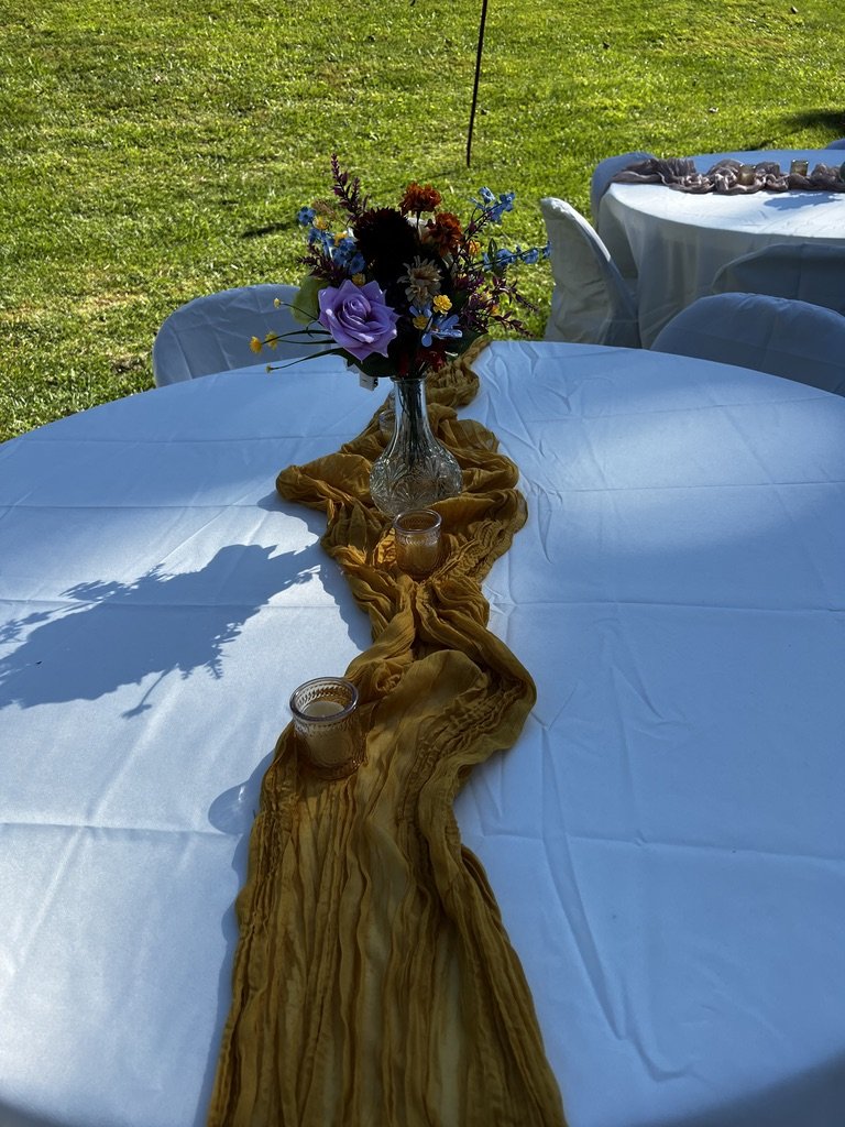Outdoor table setting with a white tablecloth, yellow table runner, and a floral centerpiece in a glass vase. Two small candles are placed on the table. The background shows more tables set on green grass.