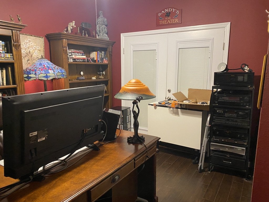 Home office with red walls, wooden desk with a monitor, a decorative lamp, bookshelves, a stereo system, and a "Home Theater" sign above a door.