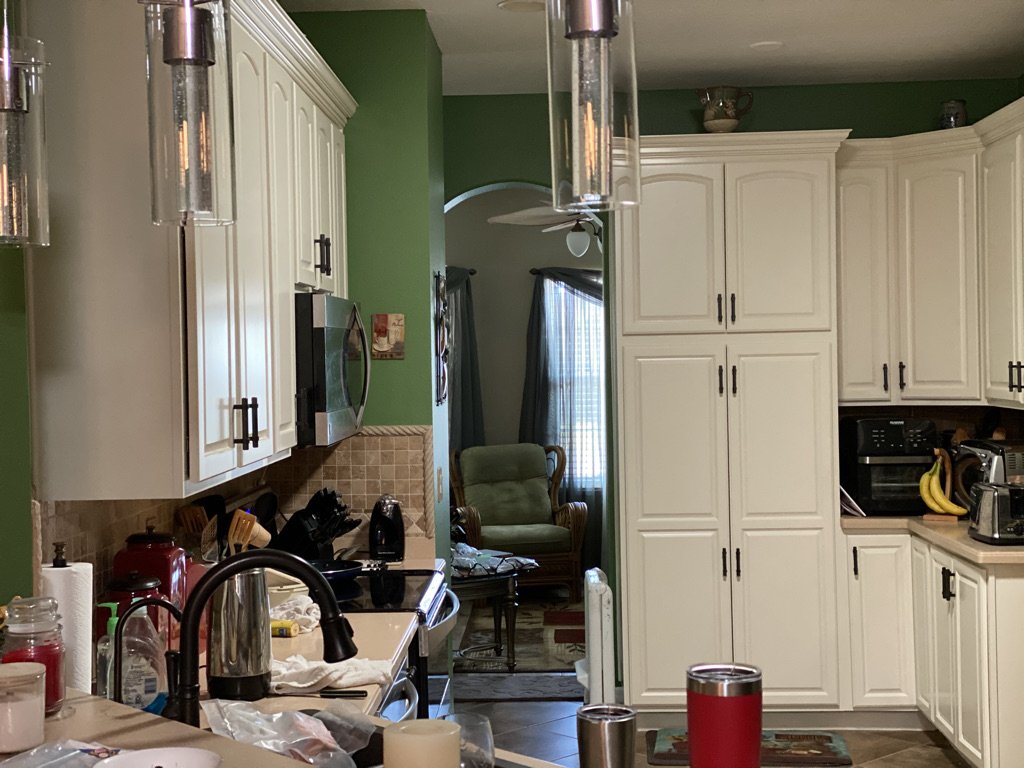 A kitchen with white cabinets, green walls, and various appliances including a microwave and toaster. The kitchen counter has a sink, dishes, and a red tumbler. Bananas are on the counter near the cabinets. In the background, an archway leads to a ro