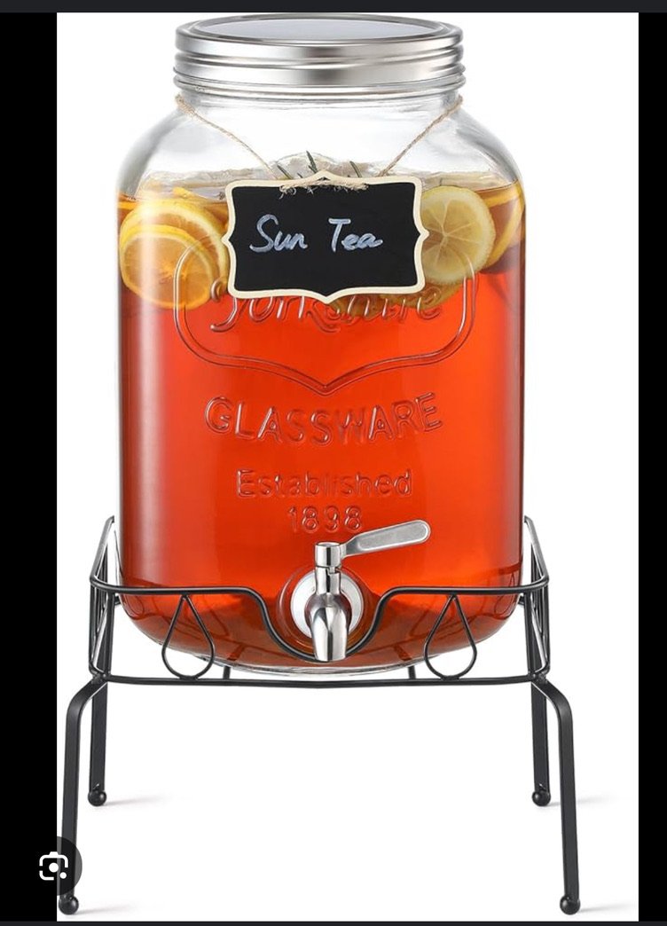 Glass beverage dispenser with sun tea and lemon slices, labeled with a chalkboard tag, on a metal stand.