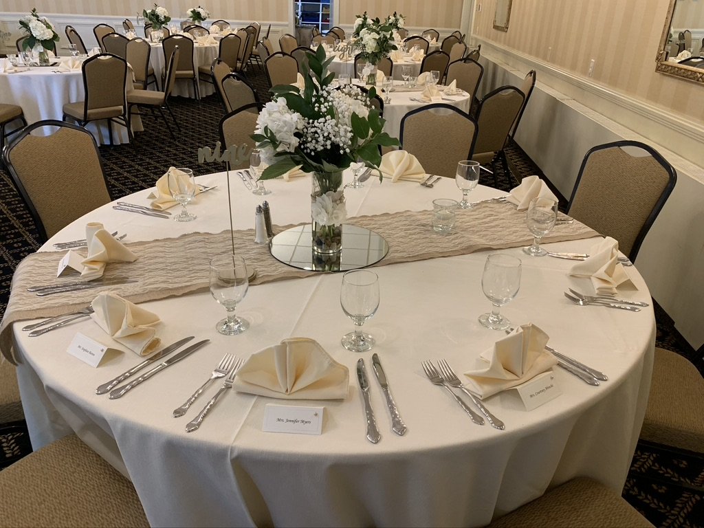Elegant banquet hall setup with round tables, white tablecloths, neatly folded napkins, silverware, and glasses. Each table has a floral centerpiece with white flowers and greenery. Chairs are arranged around the tables, and place cards are set for g