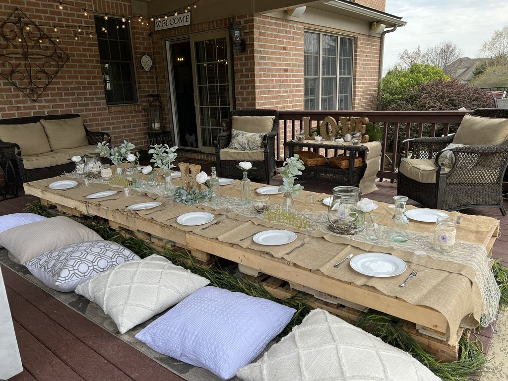 Outdoor rustic dining setup with wooden pallet table, white plates, glass jars, and floral decorations. Cushions and pillows serve as seating on a deck with wicker furniture and a "WELCOME" sign hanging on brick wall.