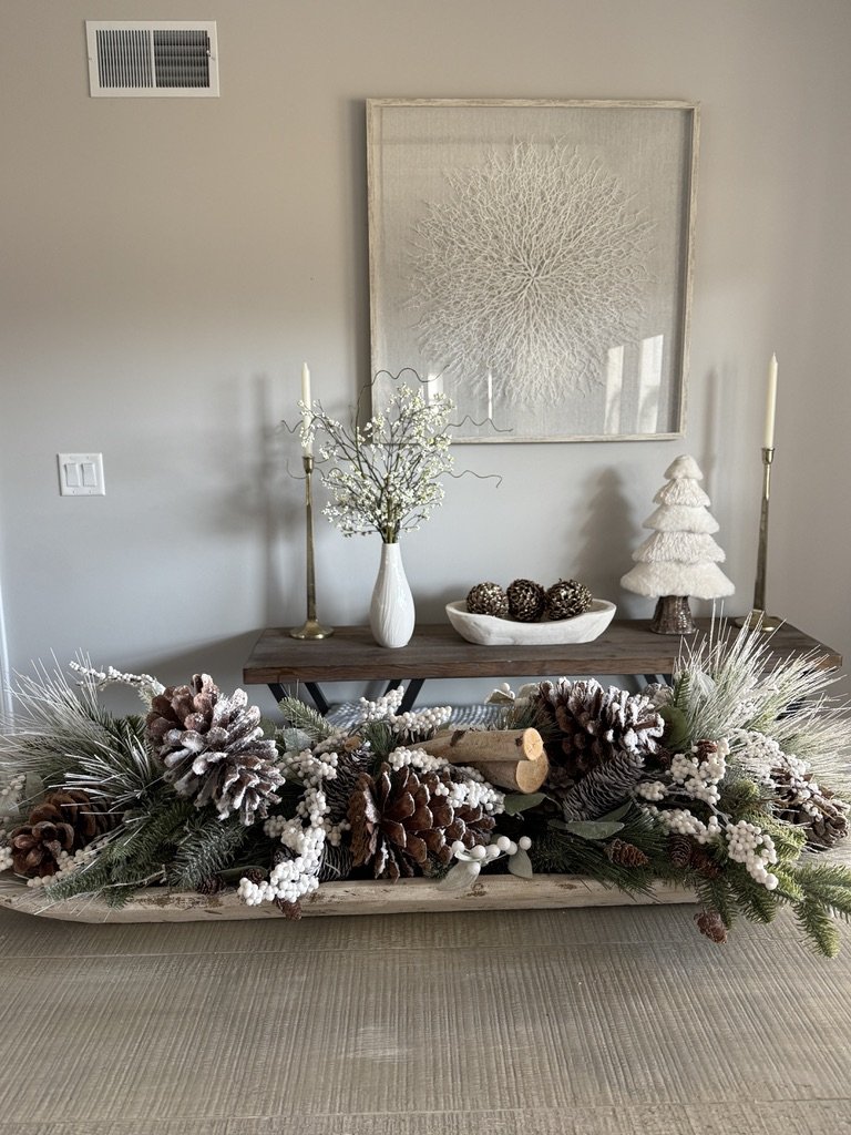 Winter-themed home decor arrangement with pinecones, greenery, and white accents on a table; background features minimalist art and decorative items.