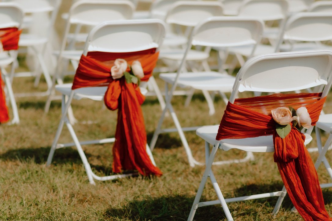 White folding chairs with orange sashes and artificial flowers on a grassy field, likely arranged for an outdoor event.